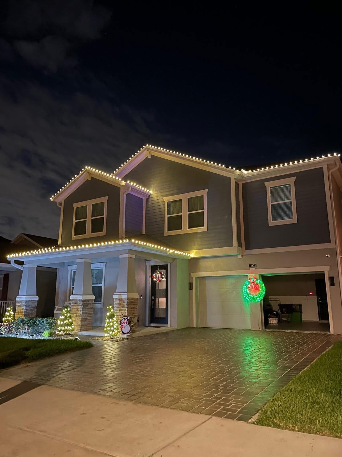 Two-story craftsman home with warm white roofline lights, lit mini trees and a wreath on the garage